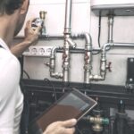 A plumber holding a tablet and inspecting water heater pipes in the boiler room