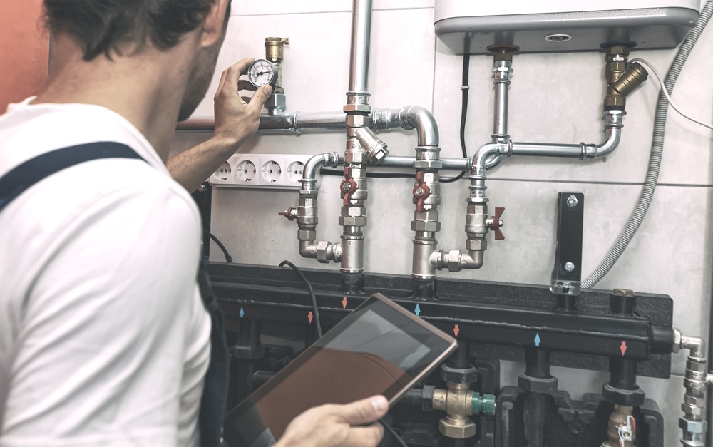 A plumber holding a tablet and inspecting water heater pipes in the boiler room