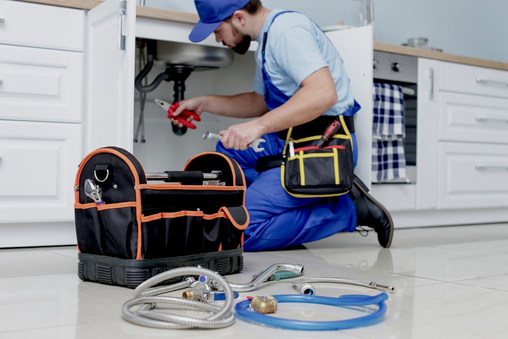 A plumber in blue overalls checking pipes under the sink with a black and orange tool box in the foreground